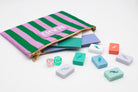 A striped zip pouch in pink and green with a gold zipper, shown open with several pastel game pieces and dice scattered in front on a white background.