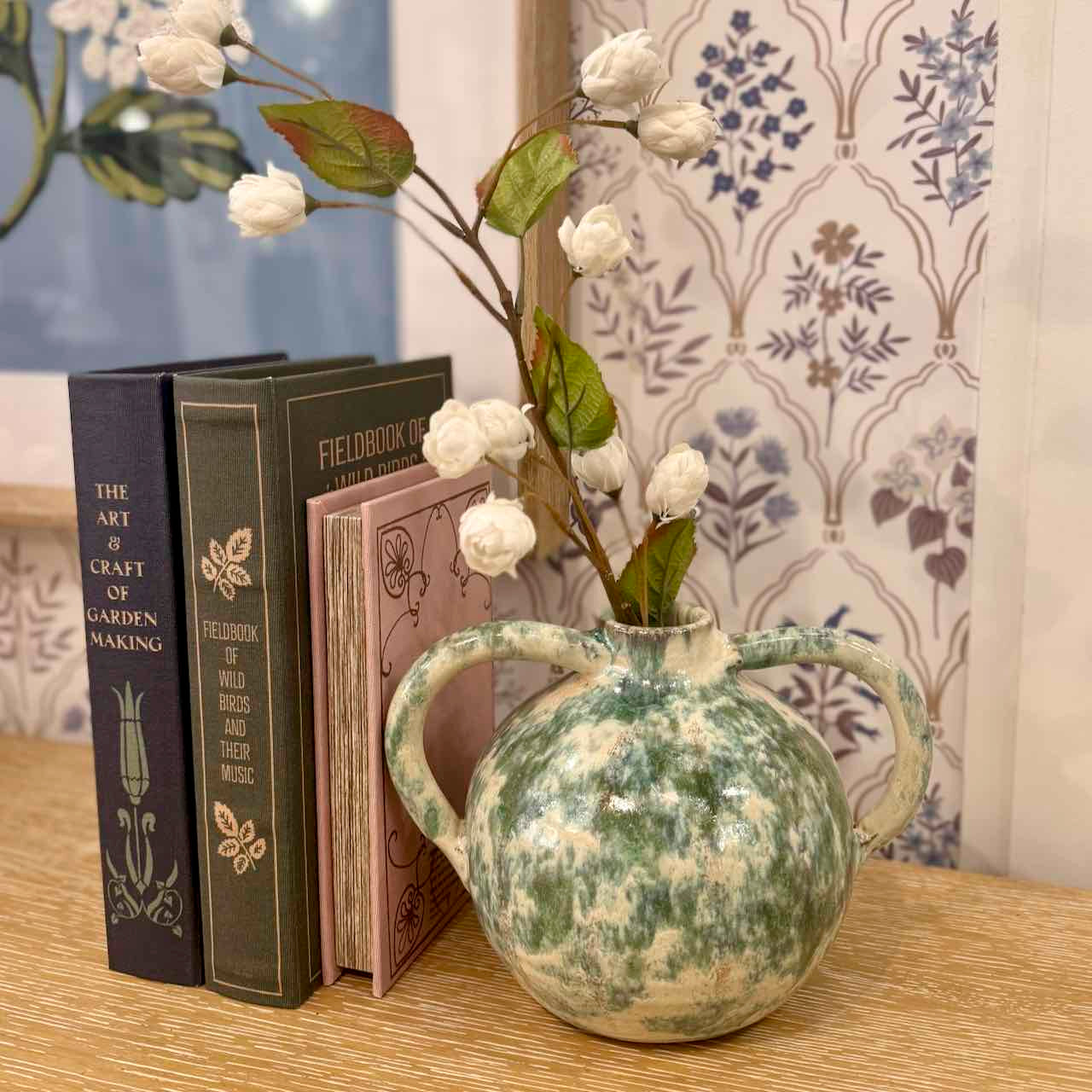 Decorative vase with white flowers on a table next to books, with a floral-patterned curtain in the background.