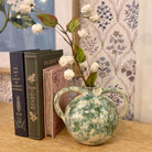 Decorative vase with white flowers on a table next to books, with a floral-patterned curtain in the background.