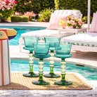 Set of turquoise glass goblets on a poolside table with lounge chairs and flowers in the background.