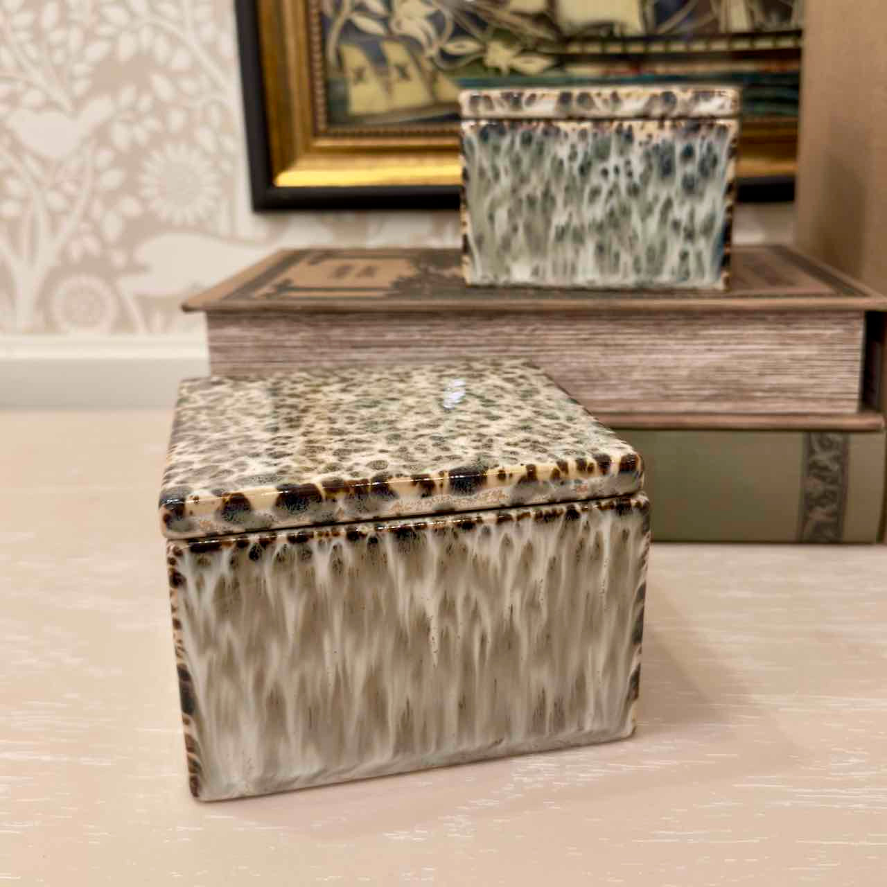 Decorative box with a patterned lid on a surface, with books and a framed picture in the background.