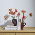 Two small dark brown ceramic pitchers with loop handles, each holding a bouquet of pink and peach carnations, styled on a light wood cabinet with books against a plain light gray wall.