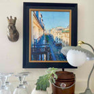 Framed painting of a cityscape with a decorative wall piece and lamp in the foreground.