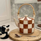 Checkered ceramic teapot with wooden handle on a round wooden board, accompanied by a cow-shaped salt shaker and spoon.
