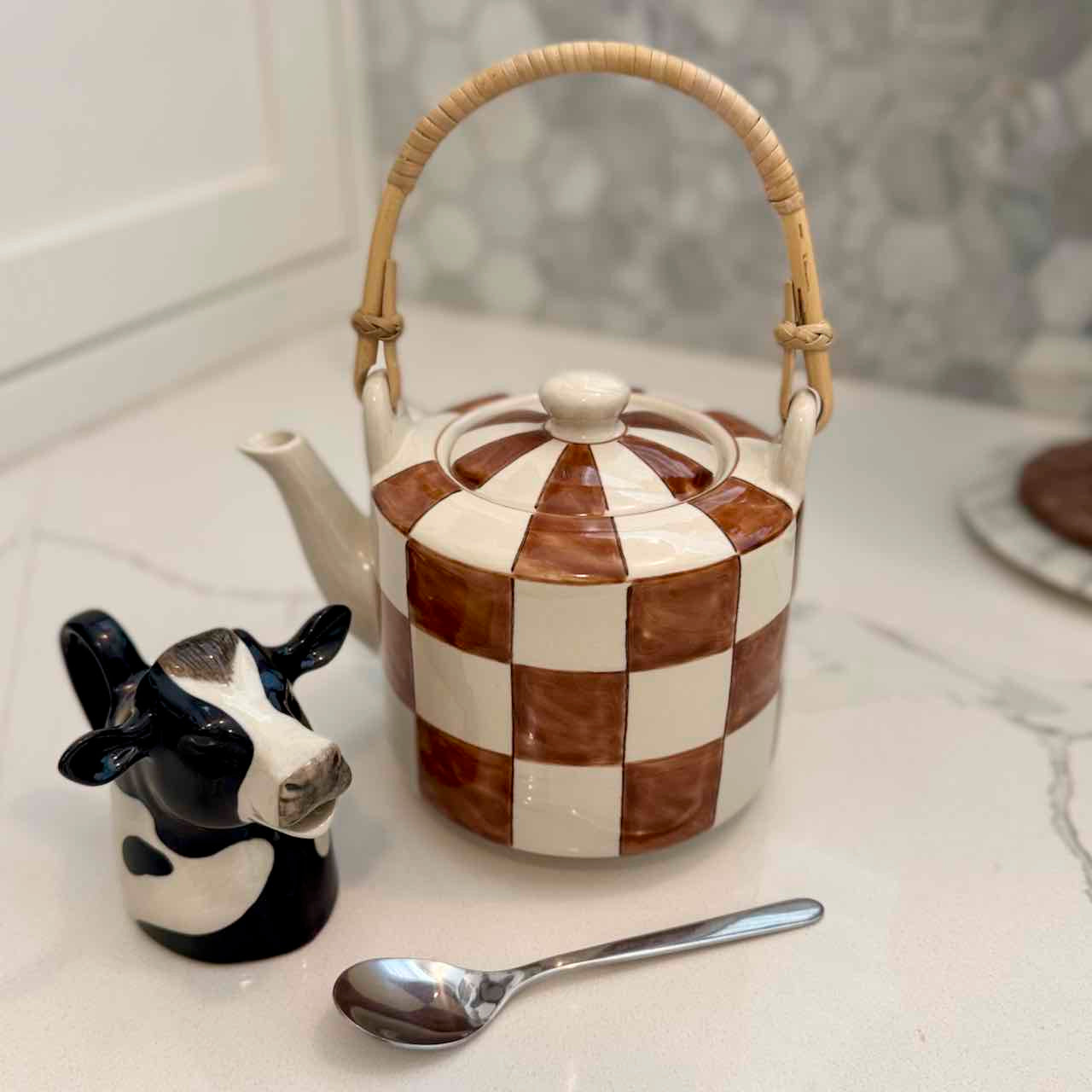 Checkered ceramic teapot with a cow-shaped salt shaker and spoon on a marble surface.