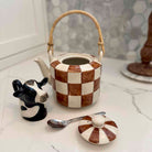 Checkered ceramic teapot with a cow-shaped salt shaker and spoon on a marble surface.