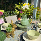 Decorative table setting with green vase, gold rabbit, and colorful flowers outdoors.