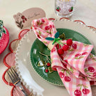 Table setting with green plate, pink napkin with cherry design, and decorative elements.