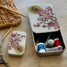 A small rectangular metal sewing kit tin with a floral lid design, shown open and closed on a wooden surface. The open tin contains thread spools, scissors, and other sewing tools.