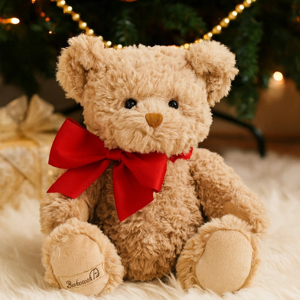 Teddy bear with a red bow sitting on a soft surface with a blurred Christmas tree in the background.