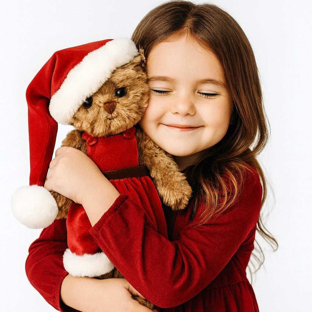 Child in a red dress holding a teddy bear wearing a Santa hat on a white background
