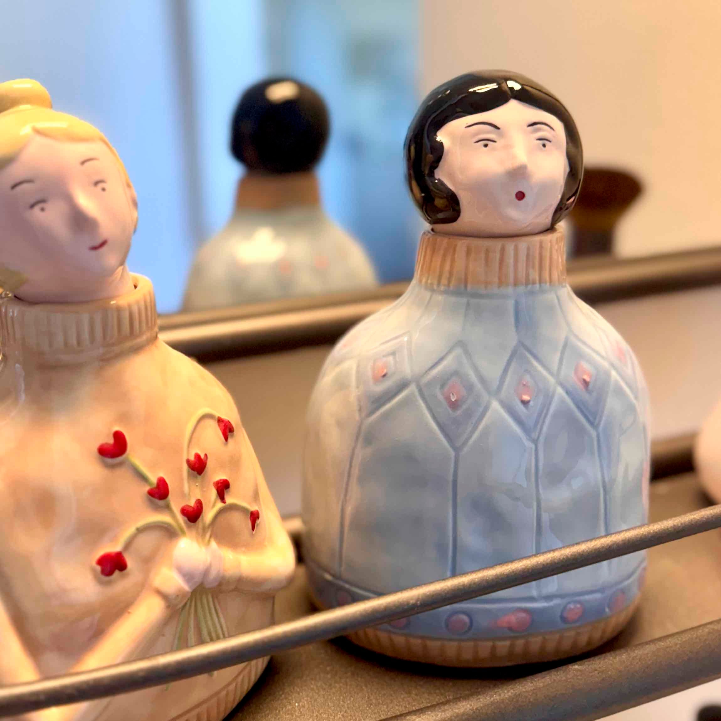Two ceramic figurines with decorative clothing and accessories on a reflective surface.