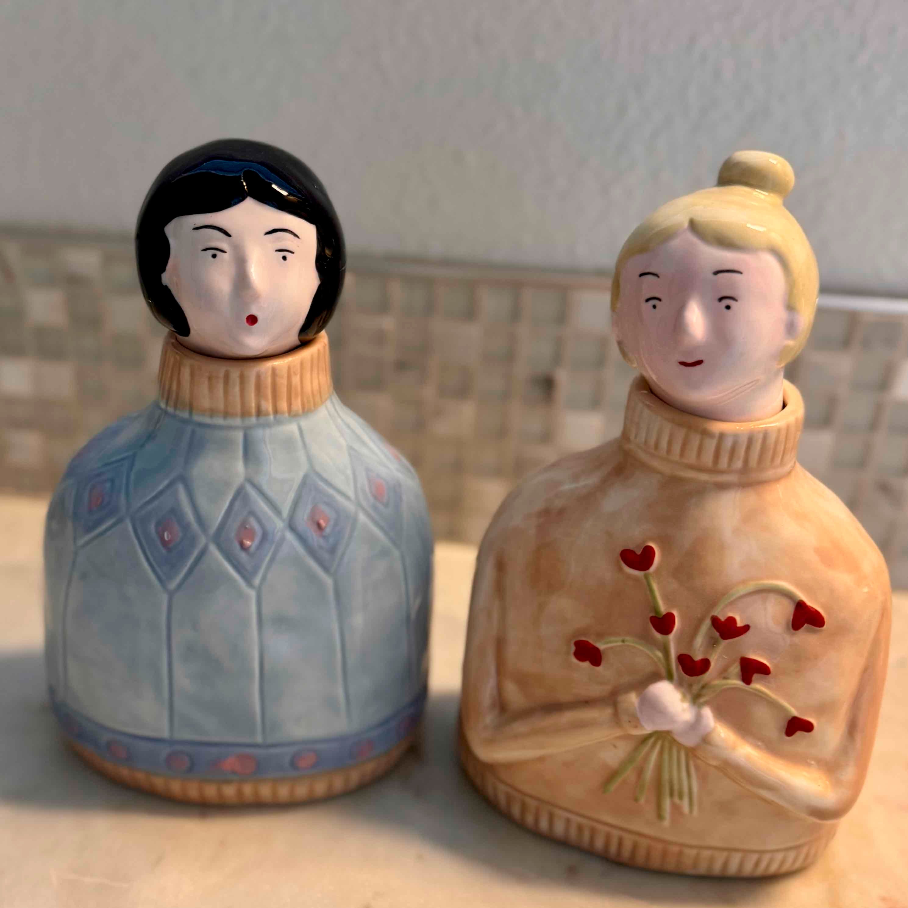Two ceramic figurines with stylized hair and clothing, one holding flowers, on a neutral background.