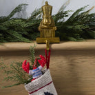 A brass nutcracker-shaped Christmas stocking hanger hanging on a wooden mantel, with a knitted Christmas stocking and green garland below.
