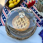 A glass dome cover with a metal latch lid sits on a woven wicker basket tray, holding a baked pie. The scene is styled on a white tablecloth with a patriotic red, white, and blue star-patterned cloth underneath, alongside lemons in a basket and a candle holder.