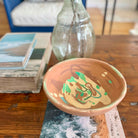 A handmade terracotta clay bowl with abstract painted patterns in green, brown, and beige, displayed on a wooden table next to stacked books and a glass vase.