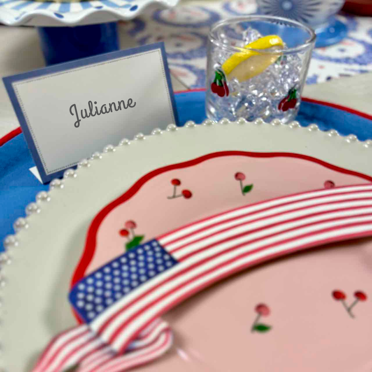 Decorative plate with American flag design, cherry illustrations, and a glass with lemon slice on a patterned tablecloth.