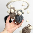 Hand holding a small sheep toy with a larger sheep toy and white beads in the background.