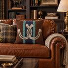 A brown leather sofa in a rustic living room setting, featuring a central decorative throw pillow with a rope-style horseshoe design over navy and teal vertical stripes. The sofa is styled with a plaid accent pillow and a cream knit throw blanket.