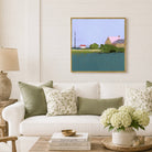 A framed landscape art print hangs on a white shiplap wall above a white sofa with green and floral throw pillows. The painting depicts a rural scene with a house, trees, and a blue-green body of water under a pale sky.