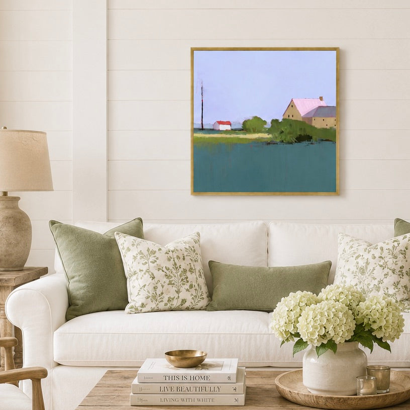 A framed landscape art print hangs on a white shiplap wall above a white sofa with green and floral throw pillows. The painting depicts a rural scene with a house, trees, and a blue-green body of water under a pale sky.