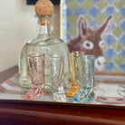 Set of four cowboy boot-shaped shot glasses in front of a bottle on a table with a checkered cloth and rabbit figurine in the background.