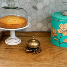 A brass spider-shaped bell on a wooden table, styled with a cake under a glass cloche on the left and a turquoise tin canister on the right, set against a hexagonal tile backsplash.