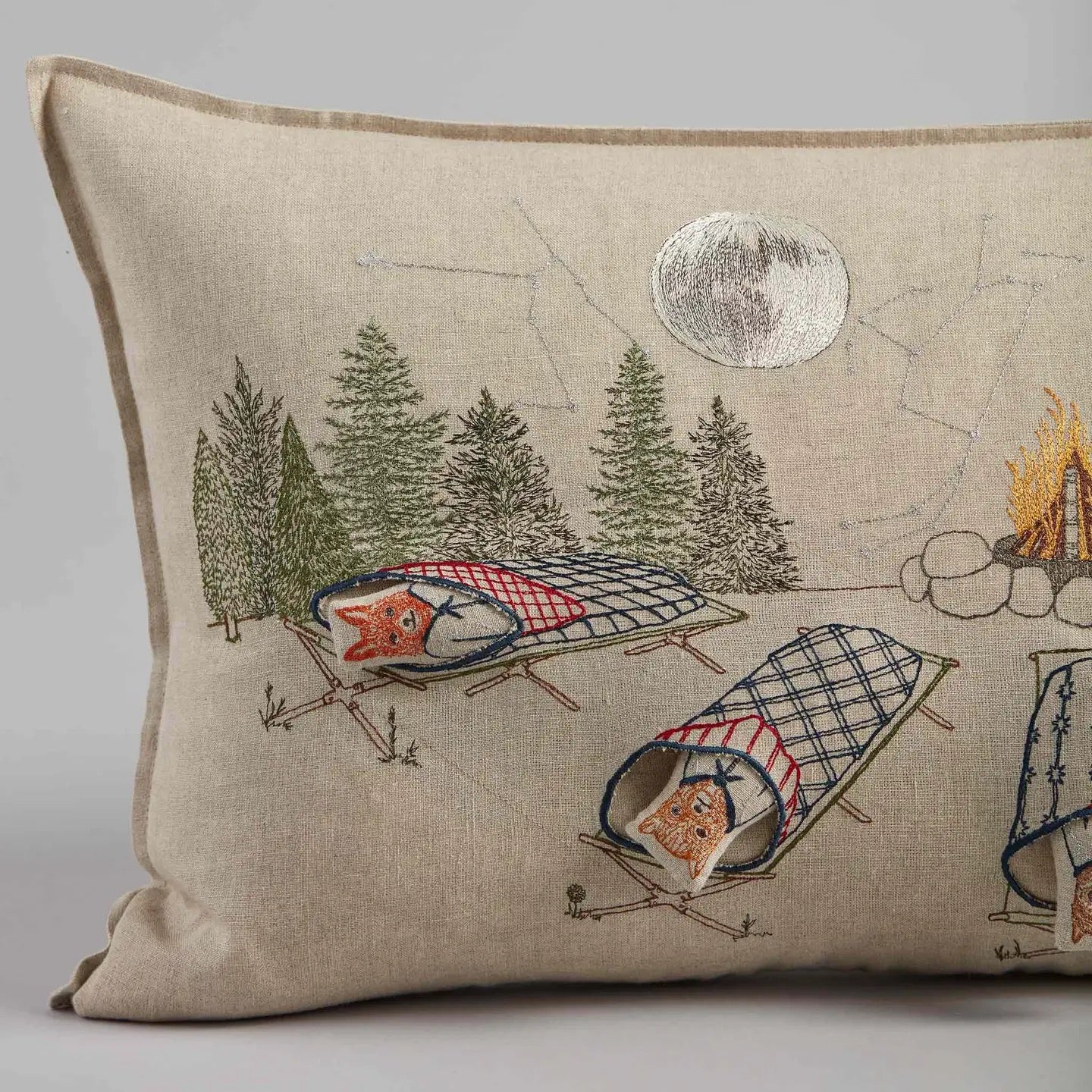 Decorative pillow with embroidered design of foxes in camping chairs by a campfire.