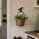 A woven wicker basket with a dark metal bird-shaped hook hanging on a beige-and-white gingham wall in a home entryway. The basket is filled with green plants and sits above white beadboard paneling, with a wooden bench and gray door visible nearby.