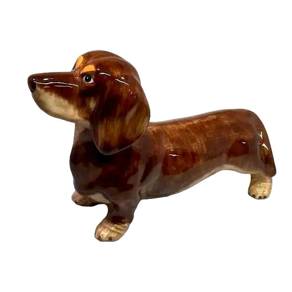 A glossy ceramic dachshund figurine in shades of brown with cream-colored paws, muzzle, and eyebrows, set against a plain light gray background.