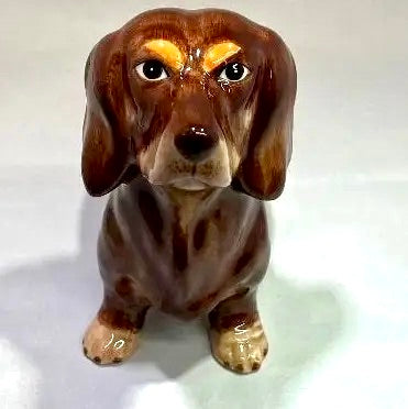 A glossy ceramic dachshund figurine in shades of brown with cream and tan accents, photographed against a plain light background.