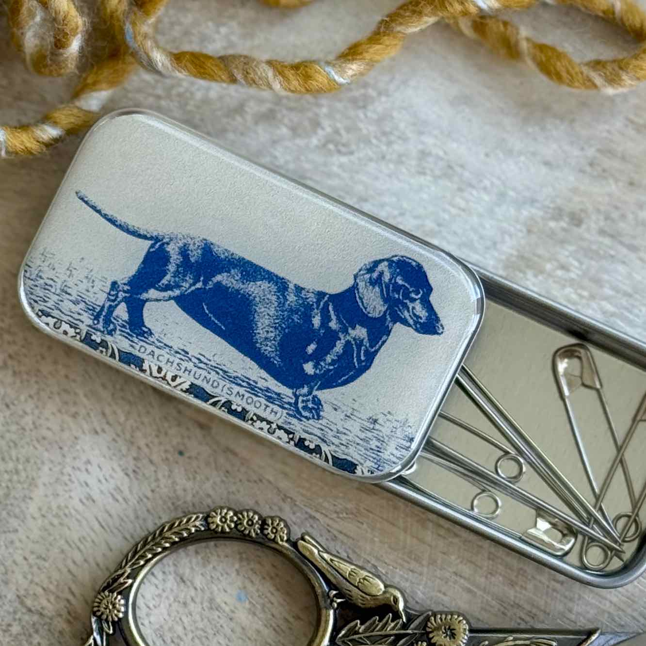 Small metal tin with a blue dachshund illustration on a textured surface with scissors and a decorative item.