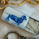 Small metal tin with a blue dachshund illustration on a textured surface with scissors and a decorative item.