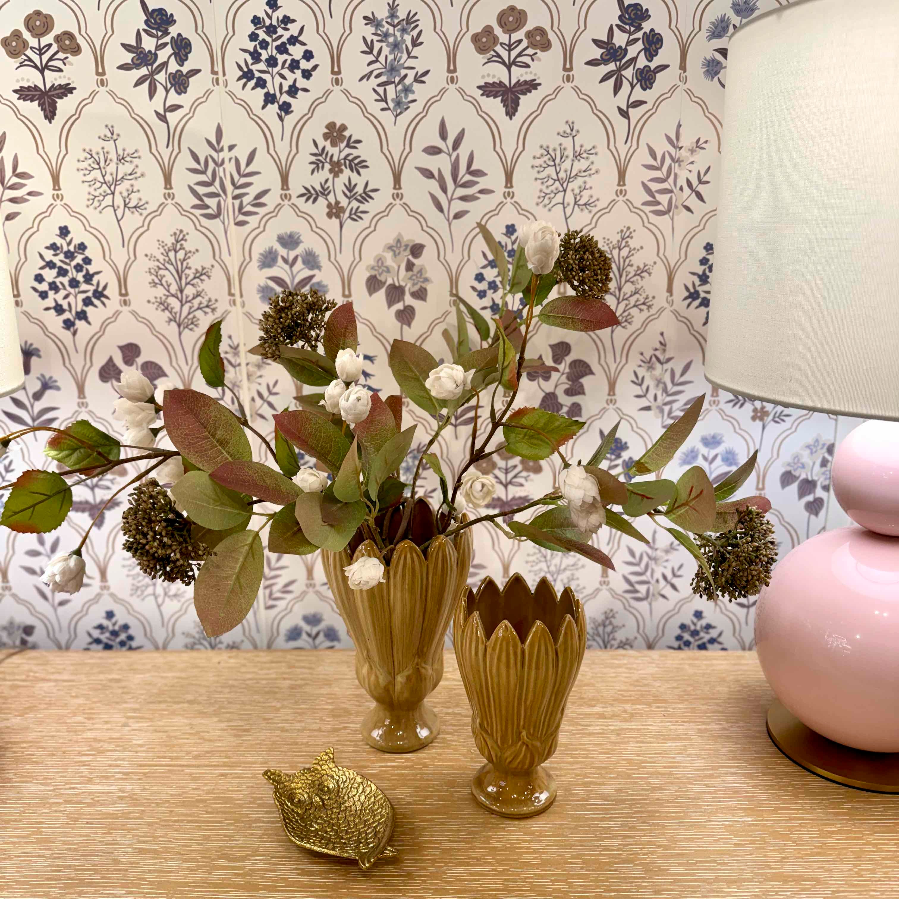 Decorative setup with floral arrangement, gold vase, gold leaf, and pink lamp against a patterned wallpaper.