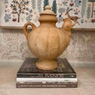 Decorative urn shaped like a chicken on top of books with a patterned wall in the background
