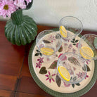 Three glasses with lemon slices on a decorative plate with floral patterns, next to a green vase and pink flowers.