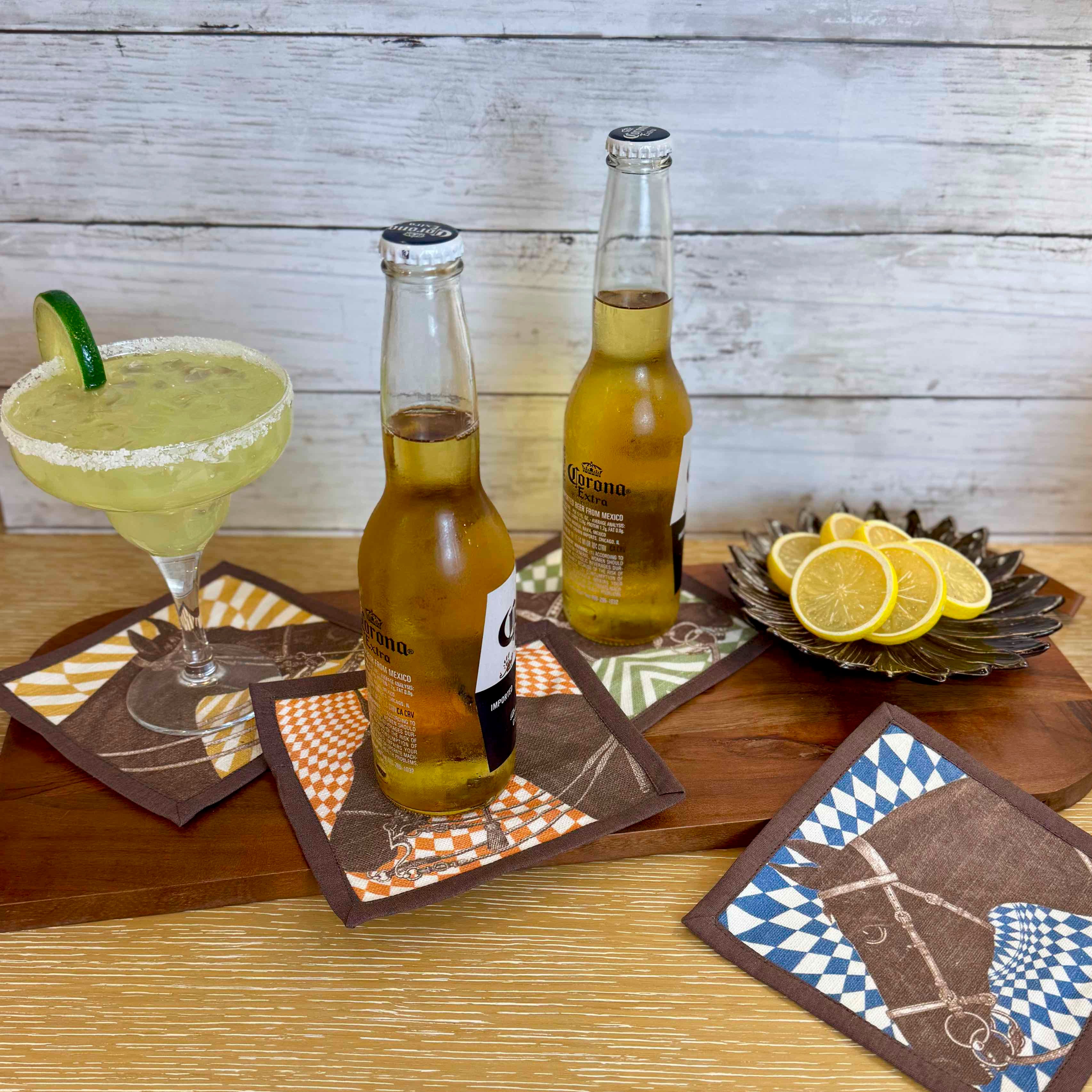 Two bottles of beer, a margarita, and lemon slices on a wooden table with patterned coasters.