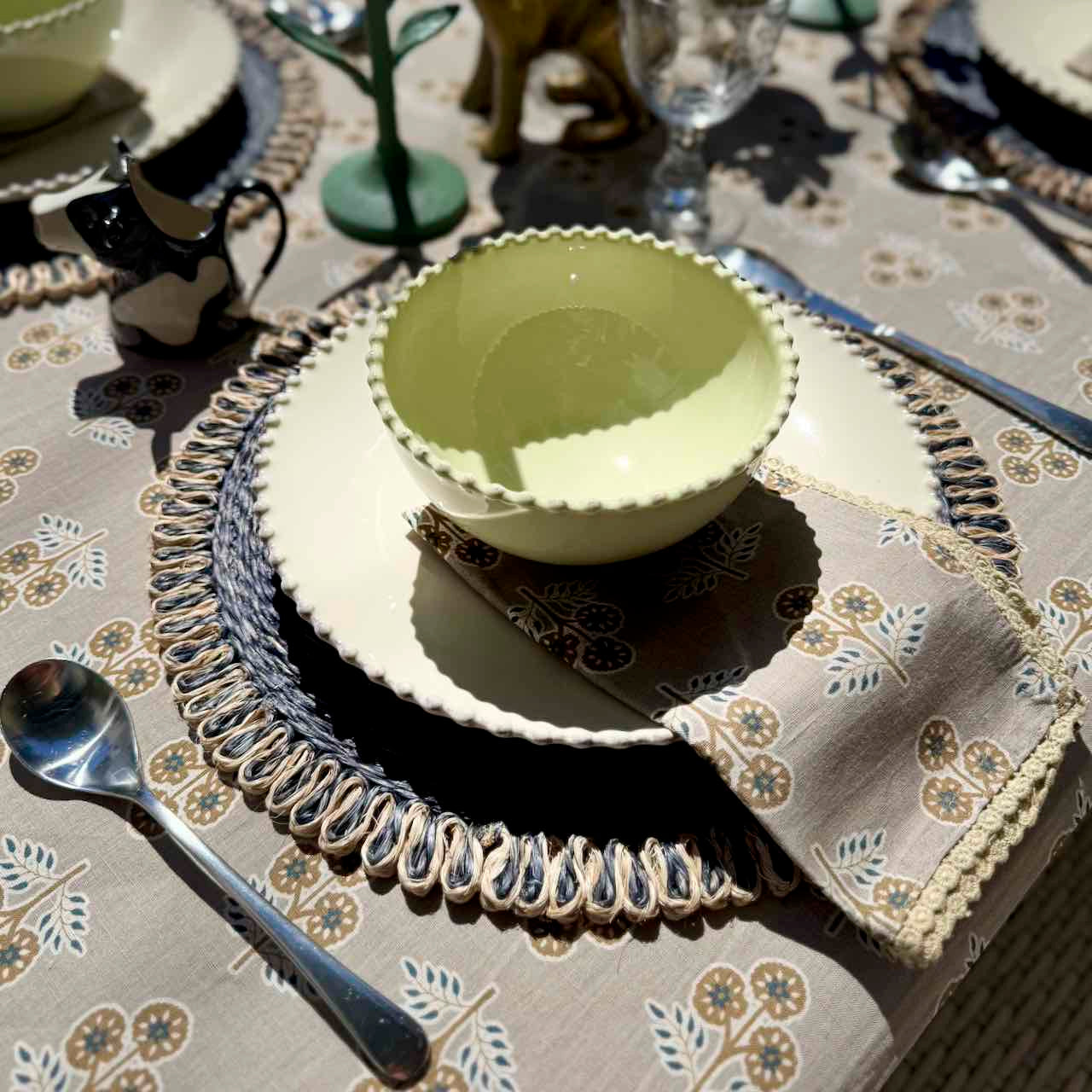Set table with green bowl, silver spoon, and patterned napkin on a floral tablecloth.