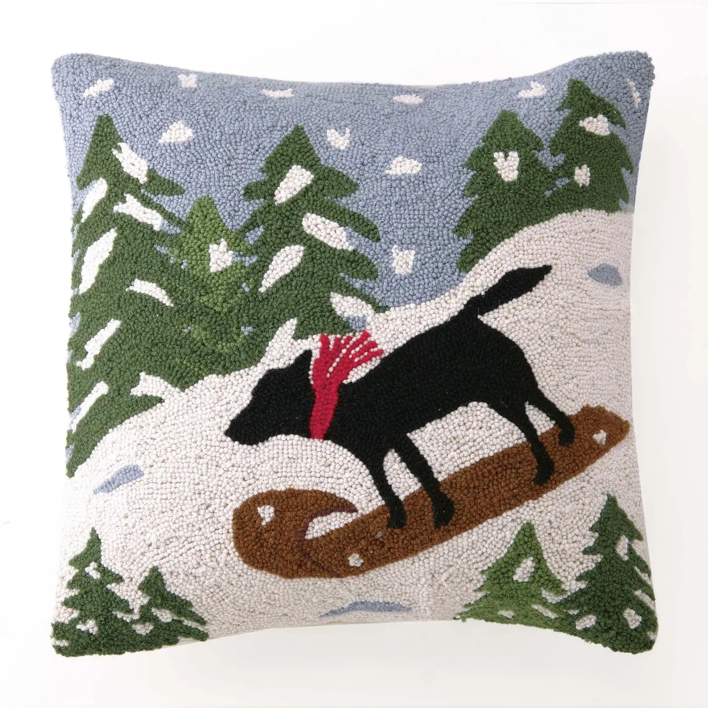 A square decorative throw pillow featuring a textured, tufted winter landscape design with a black dog on a brown sled, green pine trees, and a light blue sky.