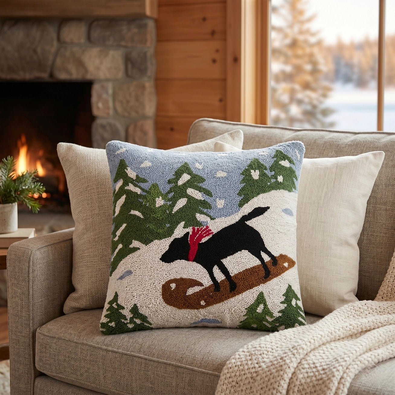 A square decorative throw pillow featuring a winter landscape design with pine trees, a black dog on a snowboard, and a red scarf, styled on a beige sofa in a cozy living room setting.
