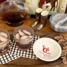 A rustic tabletop scene with two glasses of iced tea, a glass carafe, a white plate with a red squirrel logo and the words 'Don't lick my nose,' a marble mortar and pestle, silver boot-shaped cups, and decorative background objects on a wooden surface.