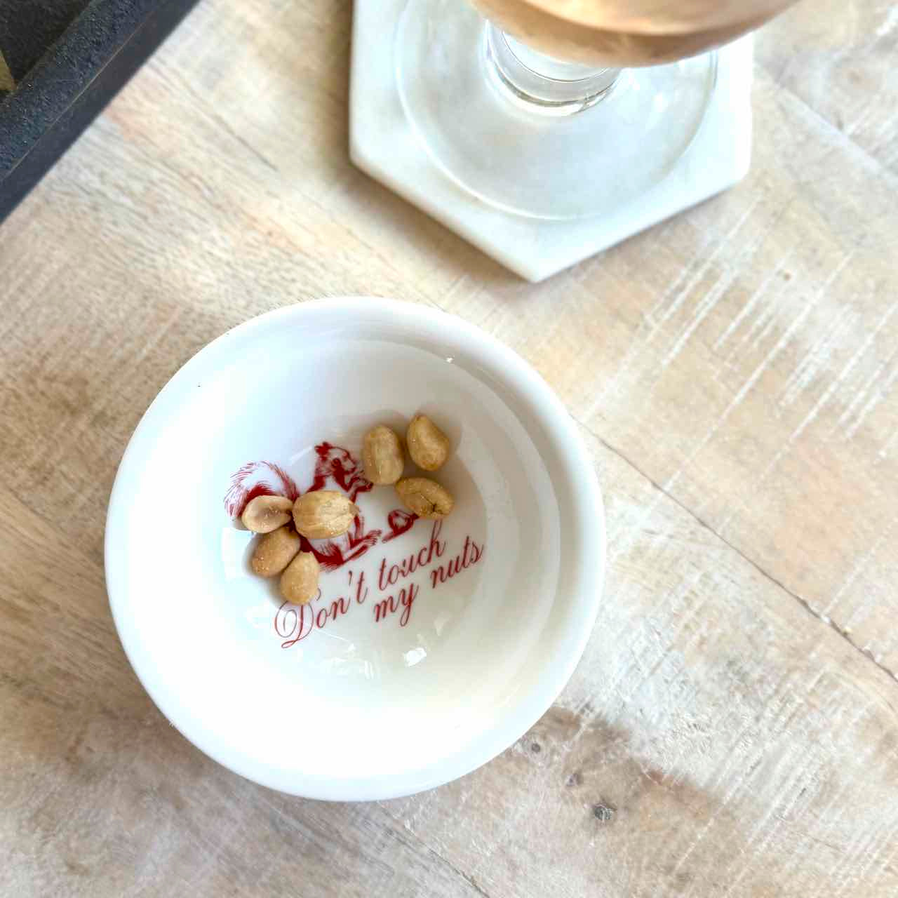 A small white ceramic bowl with the phrase 'Don't touch my nuts' printed in red script, containing a few peanuts, placed on a light wood tabletop.