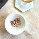 A small white ceramic bowl with the phrase 'Don't touch my nuts' printed in red script, containing a few peanuts, placed on a light wood tabletop.