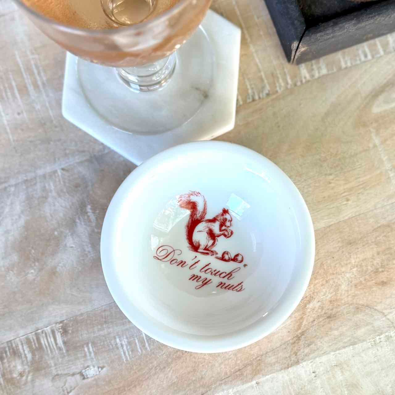 A small white ceramic dish with a red printed squirrel and the phrase 'Don't touch my nuts' on a wooden tabletop, with part of a glass and a dark rectangular object visible in the background.