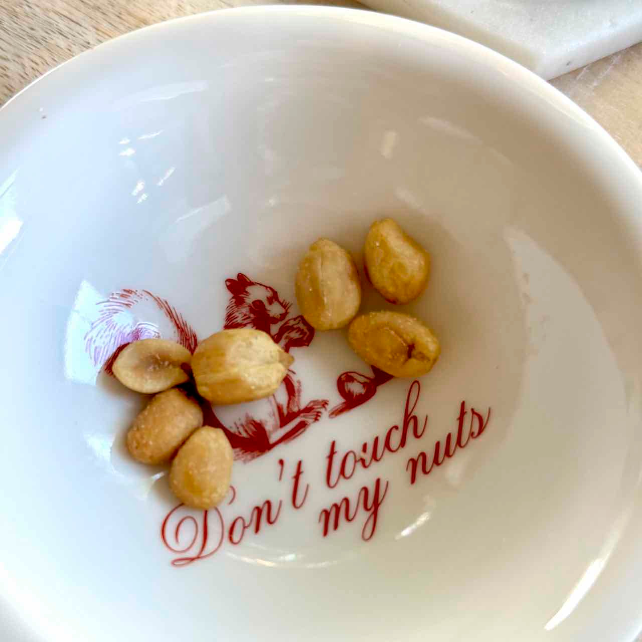 A white ceramic bowl printed with a red cartoon nut and the phrase 'Don't touch my nuts' in red script, containing several roasted peanuts inside.