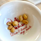 A white ceramic bowl printed with a red cartoon nut and the phrase 'Don't touch my nuts' in red script, containing several roasted peanuts inside.