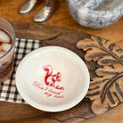 A white ceramic bowl with a red squirrel graphic and the text 'Don't touch my nuts' inside, placed on a carved wooden board next to a glass of iced tea.