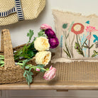 Decorative setting with a straw hat, basket of flowers, and embroidered pillow on a wooden surface.