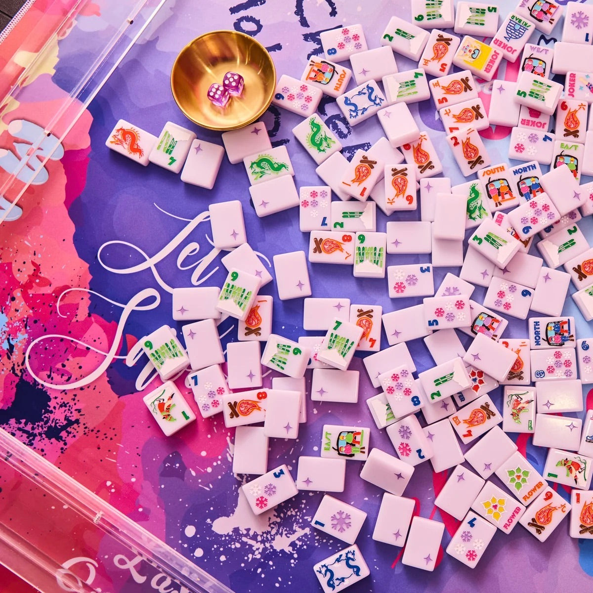 A close-up overhead view of many small white mahjong tiles scattered across a colorful purple and pink game board, with a small gold bowl holding two pink dice. The scene is styled as a product shot with no person or real environment visible.