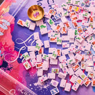 A close-up overhead view of many small white mahjong tiles scattered across a colorful purple and pink game board, with a small gold bowl holding two pink dice. The scene is styled as a product shot with no person or real environment visible.